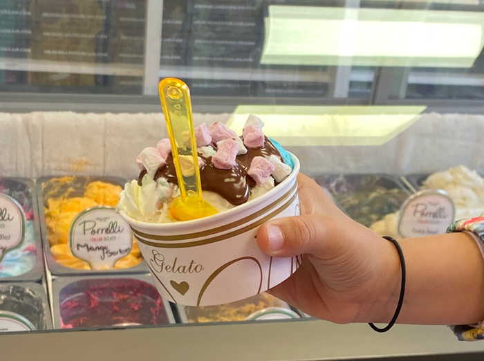 Hand holding a cup of Pirrelli Gelato topped with whipped cream, chocolate sauce, 
and mini marshmallows, purchased at Candy Cream Bathgate