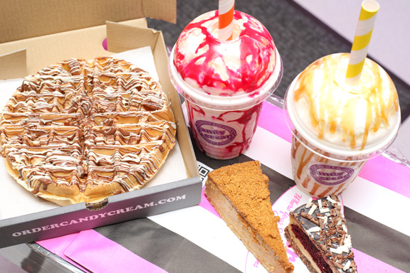Gourmet milkshake drizzled with red syrup, standing behind slices of chocolate, 
Biscoff, and fruit cheesecake from Candy Cream Bathgate